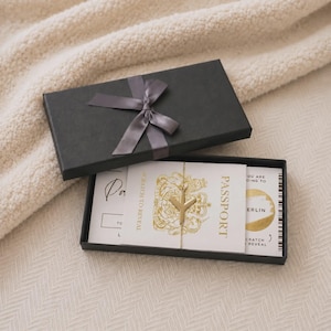 May include: A black rectangular gift box with a grey ribbon, containing a white card printed with the word PASSPORT, a gold aeroplane charm, and a scratch-to-reveal circle with the text BERLIN. The card features a gold crest and text.