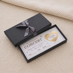 Könnte beinhalten: Eine schwarze rechteckige Geschenkbox mit einer grauen Satinschleife, die eine weiße Konzertticket-Karte enthält. Die Karte zeigt den Text "YOU ARE GOING TO A CONCERT", "Happy Sweet 16 Emily! Love, Mom & Dad", "DATE OCTOBER 18TH 2026", "TIME 07:00 PM", "VENUE HARD ROCK STADIUM" und ein goldenes Herz-Rubbelfeld mit dem Aufdruck "TAYLOR SWIFT ERAS TOUR" darunter.