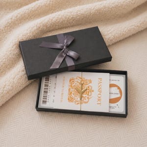 May include: A black rectangular gift box with a dark grey ribbon bow containing a travel-themed scratch-off card set. The set includes a white card with a gold aeroplane icon, the text PASSPORT, and a circular orange scratch-off area labelled BARCELONA.