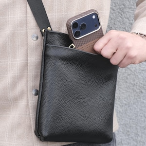 Könnte beinhalten: Schwarze Leder-Umhängetasche mit strukturierter Oberfläche. Die Tasche hat einen langen, verstellbaren Riemen und eine Tasche. Eine braune und schwarze Handyhülle ist in der Tasche sichtbar.
