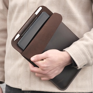 May include: A dark brown leather tablet case with a matching stylus holder. The case is open, revealing a tablet and a white stylus. The case is held by a person wearing a beige shirt.