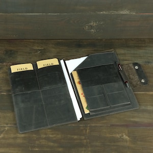 May include: A dark gray leather folio organizer featuring multiple interior pockets, card slots, and a pen loop. The left side contains two tan cards labeled FIELD, while the right side holds a notebook and a pen, secured by a snap-button flap.