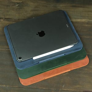 Puede incluir: Una pila de tres fundas rectangulares de cuero para tableta en azul, verde y tostado, coronada por una tableta gris oscuro con un logotipo de Apple negro y un lápiz óptico blanco que descansa a lo largo del borde inferior de la pantalla.