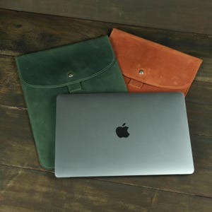 May include: A dark gray laptop with an Apple logo on the lid is positioned in front of two flat leather sleeves. One sleeve is forest green and the other is tan, both featuring a curved flap with a single silver metal snap button closure.
