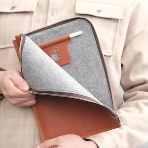 May include: A tan leather tablet sleeve featuring a grey felt interior lining, a silver metal zipper closure, and an integrated leather holder for a stylus. The interior includes a cutout detail displaying an Apple logo on a device.
