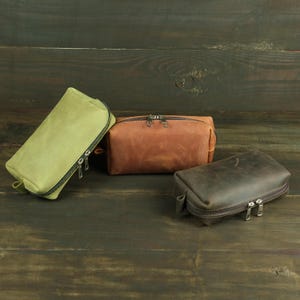 May include: Three rectangular pouches with zippered closures. One pouch is light olive green, one is medium brown, and one is dark charcoal gray. Each pouch features a smooth finish and silver-toned metal zippers with dual pull tabs.