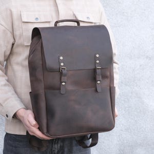 May include: A dark brown leather backpack featuring a large fold-over flap, two vertical straps with metallic buckles, a top handle, and side pockets. The bag has a smooth, matte finish and is constructed with visible stitching along the edges.