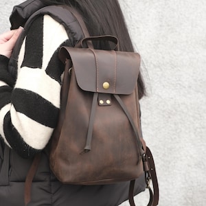 Puede incluir: Una mochila de cuero marrón con cierre de solapa y cordón. La mochila tiene un asa superior y correas ajustables. El cuero tiene un aspecto envejecido. La mochila se lleva sobre un suéter a rayas blancas y negras.