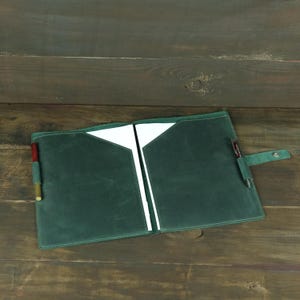 Könnte beinhalten: Ein dunkelgrüner Notizbuchumschlag aus Leder mit zwei Innentaschen für weißes Papier, einer Stiftschlaufe auf jeder Seite mit einem rot-goldenen Stift links und einem schwarzen Stift rechts sowie einem Riemen mit silbernem Druckknopfverschluss.