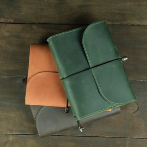 May include: Three leather journals stacked together, featuring a green, a tan, and a dark brown cover. Each journal has a rounded flap closure secured by a thin black elastic cord with a small metallic bead at the end.