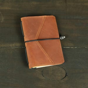 May include: A tan leather notebook cover featuring a diagonal stitched pocket on the front, secured by a black elastic band with a small silver-toned metal charm shaped like a compass rose attached to the side.