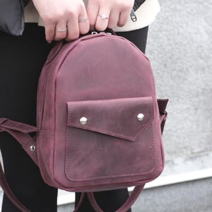 May include: A deep burgundy leather backpack featuring a rounded top, a main zippered compartment, and a front pocket with an asymmetrical flap secured by two silver-toned metal snap buttons. The bag includes adjustable shoulder straps.