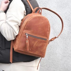 Può includere: Uno zaino in pelle color cuoio con parte superiore arrotondata, una tasca frontale con cerniera dotata di tirante con nappa in pelle abbinata e spallacci regolabili. La borsa presenta una finitura opaca liscia con grana naturale e dettagli argentati.