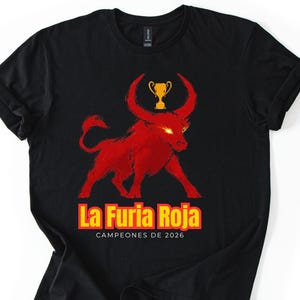 May include: A black short-sleeve t-shirt featuring a graphic of a red bull with glowing eyes and a gold trophy above it. Below the bull, the text reads La Furia Roja and CAMPEONES DE 2026 in yellow and white lettering on the front of the garment.