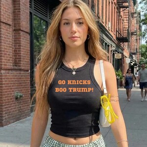 May include: A black sleeveless tank top featuring the orange text "GO KNICKS BOO TRUMP!" printed on the front. The top is paired with green and white gingham patterned pants and a white shoulder bag with yellow-rimmed sunglasses attached.