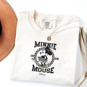 May include: Cream-coloured t-shirt with a Minnie Mouse graphic. The design includes the text "Minnie Mouse" and "1928" within a chequered circle. A gold necklace and a brown hat are also visible.
