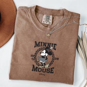 May include: A brown Comfort Colors t-shirt with a Minnie Mouse graphic. The graphic includes the text "Minnie Mouse" and "1928". A silver necklace with a pendant is draped across the shirt.