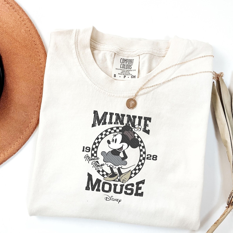 May include: Cream-coloured t-shirt with a Minnie Mouse graphic. The design includes the text "Minnie Mouse" and "1928" within a chequered circle. A gold necklace and a brown hat are also visible.