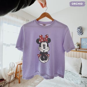 May include: A light purple t-shirt featuring a graphic of Minnie Mouse with a red polka dot bow. The t-shirt is hanging on a wooden hanger. The word "ORCHID" is in a purple rectangle in the top right corner.