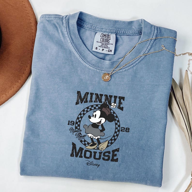 May include: A pale blue Comfort Colors t-shirt with a Minnie Mouse graphic. The design includes the text "Minnie Mouse" and the years "1928" and "19". A gold necklace with a pendant is placed on the shirt.
