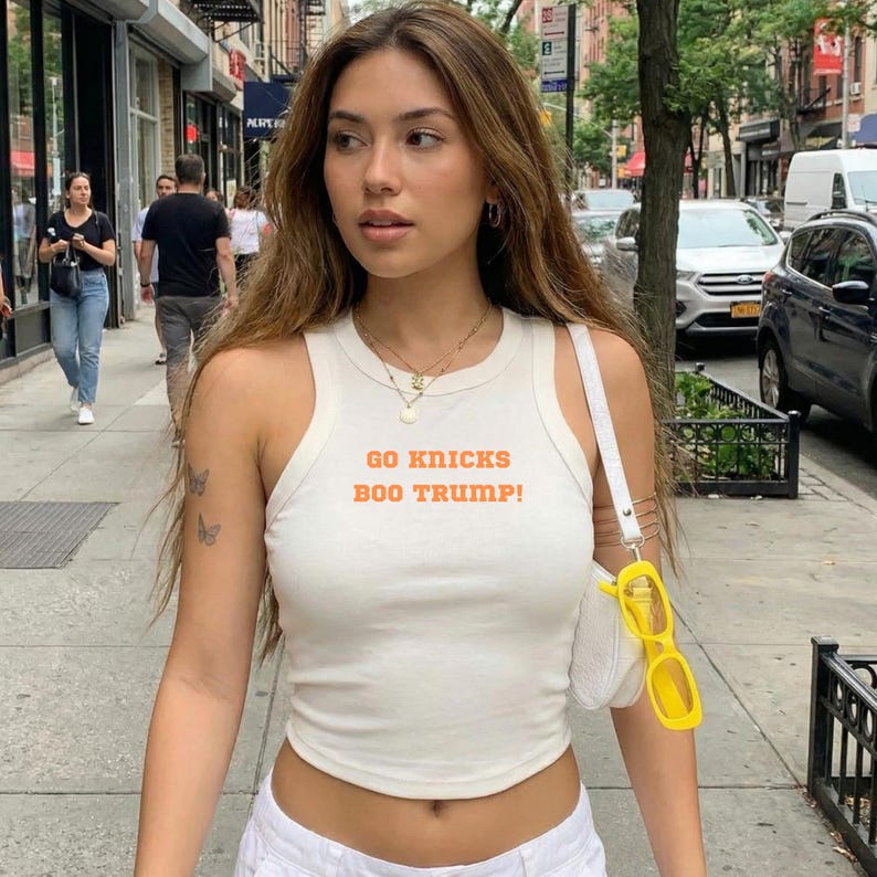 May include: A white sleeveless racerback tank top featuring the orange text GO KNICKS BOO TRUMP! printed on the chest. The garment has a round neckline and a fitted silhouette, paired with a white shoulder bag and bright yellow rectangular sunglasses.