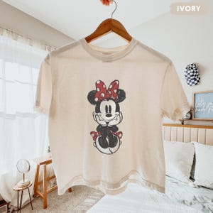 May include: Ivory-colored t-shirt featuring a distressed graphic of Minnie Mouse. The cartoon character is depicted with a red polka-dot bow, black dress, and white gloves. The shirt is hanging on a wooden hanger.