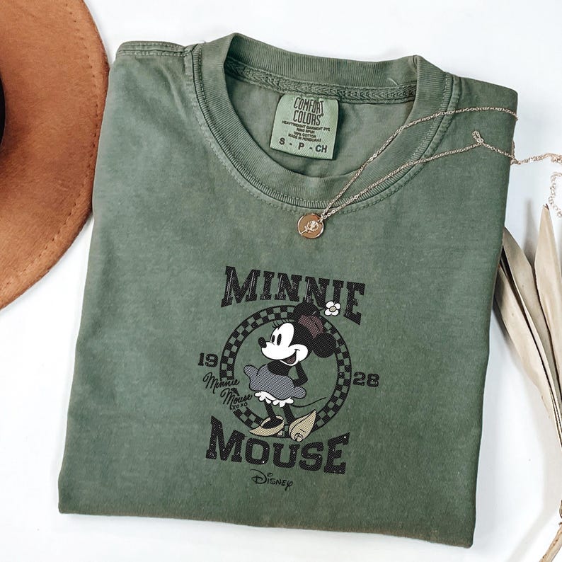 May include: An olive green t-shirt with a Minnie Mouse graphic. The design includes the text "Minnie Mouse" and the years "1928" and "1928". A gold necklace is draped across the shirt.