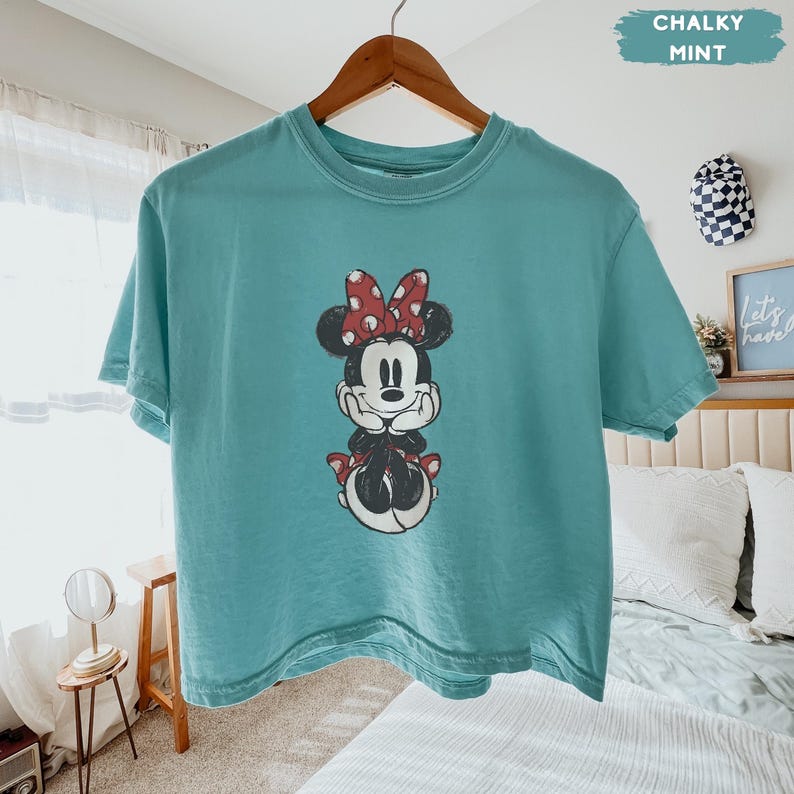 May include: A teal-colored t-shirt featuring a graphic of Minnie Mouse with a red bow and polka dots. The t-shirt is hanging on a wooden hanger. The words "Chalky Mint" are in a teal box in the upper right corner.
