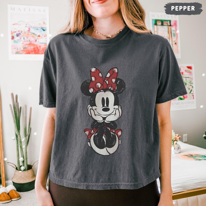 May include: A dark gray t-shirt featuring a distressed graphic of Minnie Mouse. Minnie is depicted with a red bow with white polka dots, a black dress, and red shoes. The word "PEPPER" is in the top right corner.