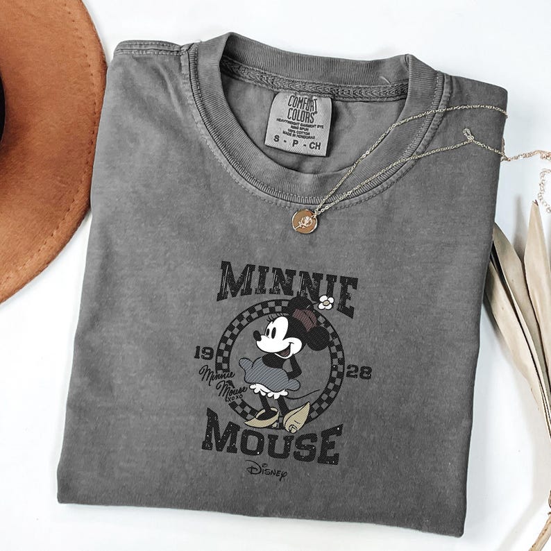 May include: A grey Comfort Colors t-shirt with a Minnie Mouse graphic. The graphic includes the text "Minnie Mouse" and "Disney". A silver necklace with a pendant is draped over the shirt. A brown hat is visible in the upper left corner.