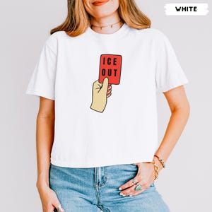 May include: A white short-sleeved t-shirt featuring a graphic of a hand holding a red rectangular card with the black text ICE OUT printed on it. The design is centered on the chest of the shirt, which has a crew neckline and a standard fit.