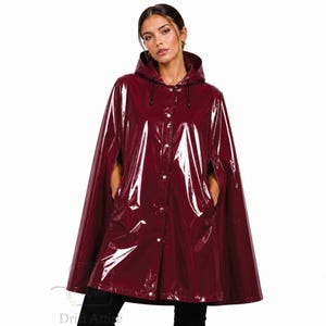 May include: A glossy, deep burgundy vinyl rain cape featuring a hood with black drawstring cords, a front button-down closure with silver-toned metal snaps, and side arm openings. The garment has a smooth, reflective finish and a loose, flowing fit.