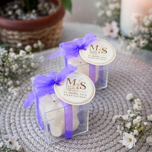 Könnte beinhalten: Zwei transparente quadratische Boxen mit weißen Kerzen, gebunden mit lila Bändern. Jede Box hat ein rundes Etikett mit den Initialen "M & S" und türkischem Text. Die Boxen stehen auf einer grauen gewebten Matte, mit Blumen und einer brennenden Kerze.