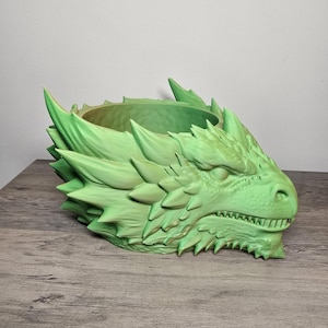 May include: A light green dragon head planter featuring detailed scales, sharp spikes along the sides, and a wide open top. The creature has a defined snout, visible teeth, and a textured surface throughout the entire structure of the vessel.