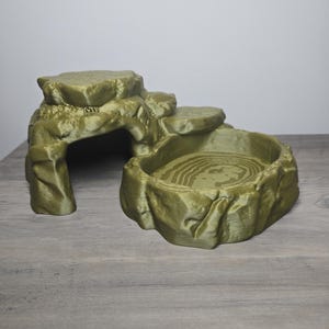 May include: An olive green reptile hide and water dish combination. The structure features a raised rock-like platform with a hollow cave entrance on the left and a shallow, oval-shaped basin with concentric ring textures on the right.