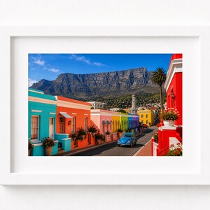 Può includere: Una stampa fotografica incorniciata che mostra una strada fiancheggiata da case dai colori vivaci in turchese, arancione, rosa e rosso. Un'auto blu percorre la strada verso una grande montagna sotto un cielo azzurro, in una cornice bianca.