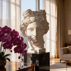 May include: A white marble bust of a classical figure sits atop a black marble pedestal. The bust is in a well-lit room with a window and curtains. A vibrant purple orchid plant is in the foreground.