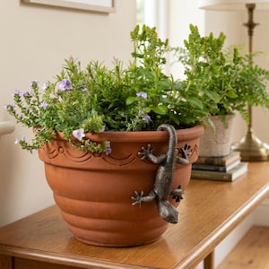 May include: A terracotta plant pot filled with green plants and purple flowers. A bronze lizard decoration clings to the side. The pot is on a wooden table, with a lamp and other plants in the background.