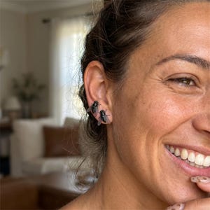 Op de afbeelding: Close-up van een oor met twee vliegvormige oorbellen. De oorbellen zijn zwart met gedetailleerde vleugels, wat een uniek accessoire creëert. De persoon glimlacht, waardoor de oorbellen worden benadrukt.