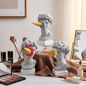 Könnte beinhalten: Drei graue Büsten-Skulpturen mit Eiswaffel-Augenersatz. Eine Waffel ist gelb mit rotem Akzent, eine andere gelb mit blauem Akzent und die dritte gelb mit cremefarbenem Akzent. Make-up-Pinsel, Lippenstifte und ein Spiegel sind ebenfalls im Bild.