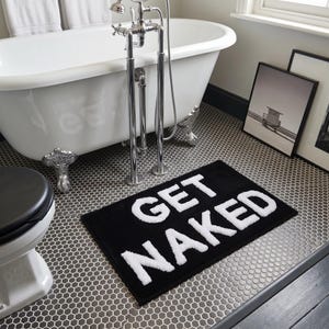 Può includere: Un tappetino da bagno rettangolare nero con le parole "GET NAKED" a grandi lettere bianche. Il tappetino è posizionato su un pavimento piastrellato grigio e bianco in un bagno, vicino a una vasca da bagno bianca.