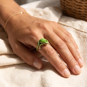May include: A green frog ring with gold detailing. The frog design wraps around the finger. The ring is made of metal and features a detailed frog design. The ring is a unique piece of jewellery.