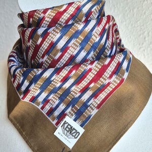May include: A square scarf featuring a central pattern of diagonal stripes in red, blue, and tan with white accents. The scarf has a solid tan border and a white label with the text KENZO in black, along with a signature in blue script embroidery.