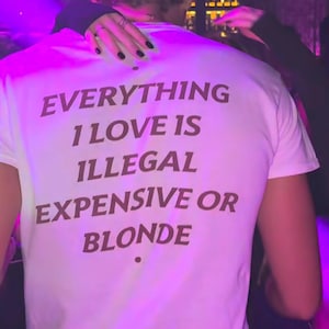 May include: A white short-sleeved t-shirt featuring dark brown text on the back that reads: EVERYTHING I LOVE IS ILLEGAL EXPENSIVE OR BLONDE. The shirt has a crew neckline and is worn in a dimly lit environment with purple ambient lighting.