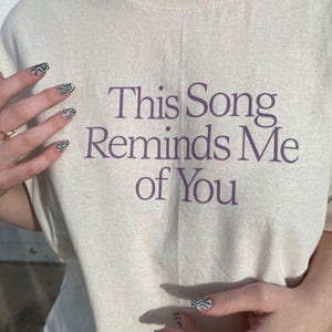May include: Cream-colored t-shirt with the text "This Song Reminds Me of You" in purple. The shirt is worn by someone with green and white patterned nails and gold rings.