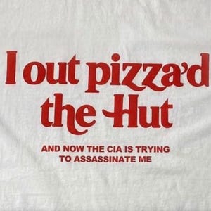 Może przedstawiać: Biała koszulka z pogrubionym czerwonym napisem I out pizza'd the Hut, a pod nim mniejszym czerwonym tekstem o treści AND NOW THE CIA IS TRYING TO ASSASSINATE ME. Napis został wydrukowany wyraźną czcionką bezszeryfową na jasnym tle materiału.