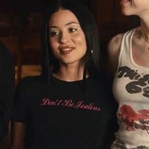 Peut inclure: Un t-shirt noir avec le texte "Don't Be Jealous" en script rose. Le t-shirt est port&eacute; par une personne aux cheveux fonc&eacute;s et maquill&eacute;e. Une autre personne est partiellement visible &agrave; droite, portant un d&eacute;bardeur blanc.