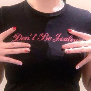 Peut inclure: T-shirt noir avec le texte rose "Don't Be Jealous". Le t-shirt est de style col rond. L'image montre &eacute;galement des mains avec du vernis &agrave; ongles rouge et &agrave; motifs.