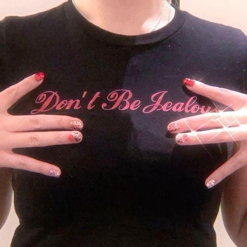 Peut inclure: T-shirt noir avec le texte rose "Don't Be Jealous". Le t-shirt est de style col rond. L'image montre &eacute;galement des mains avec du vernis &agrave; ongles rouge et &agrave; motifs.