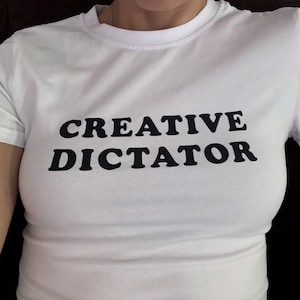 May include: A white short-sleeved t-shirt featuring the text CREATIVE DICTATOR printed in bold black capital letters across the chest. The garment has a classic crew neckline and a fitted silhouette, providing a clean and simple aesthetic.